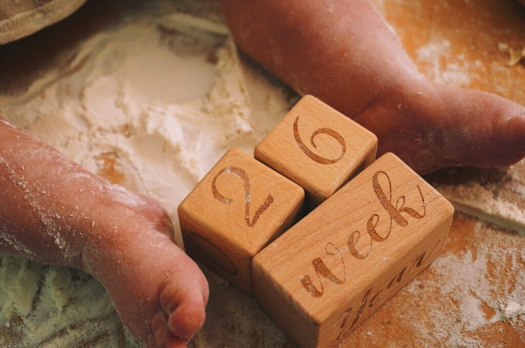 Adorable baby feet on flour-covered surface, next to wooden milestone blocks displaying 26 weeks.